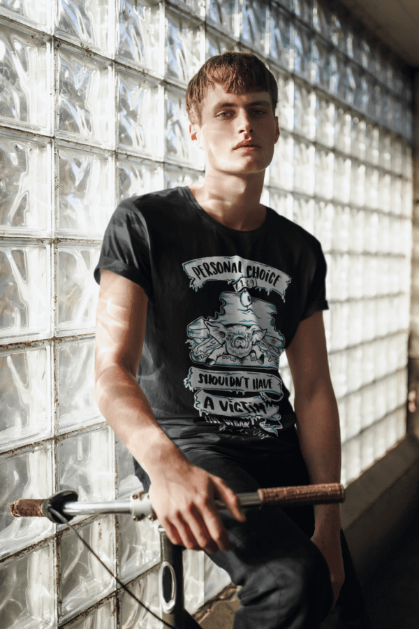 A Young male model posing with a bike and leaning against a window wearing a black organic T-shirt with illustraion of a live pig in a sandwich and the wording "personal choice shouldn't have a victim