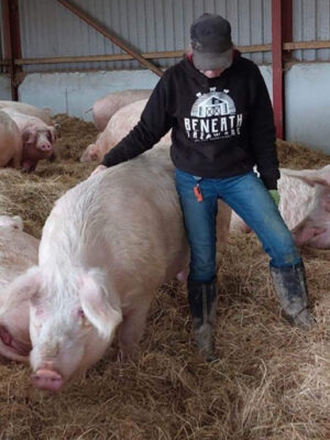 Sasha standing up stroking a pig wearing a black hoodie with white print & hot pink trims. By Viva La Vegan. Fund raising for Beneath The Wood Sanctuary