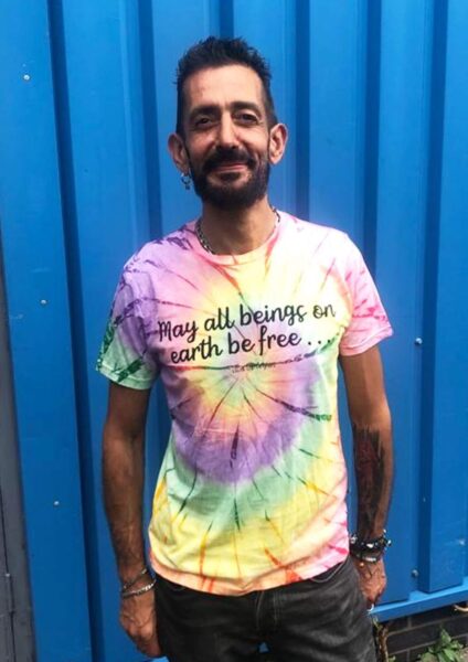 Dark haired man wearing a rainbow tie dye tshirt with the slogan May all Beings Be Free print on the chest by eco ethical brand Viva La Vegan