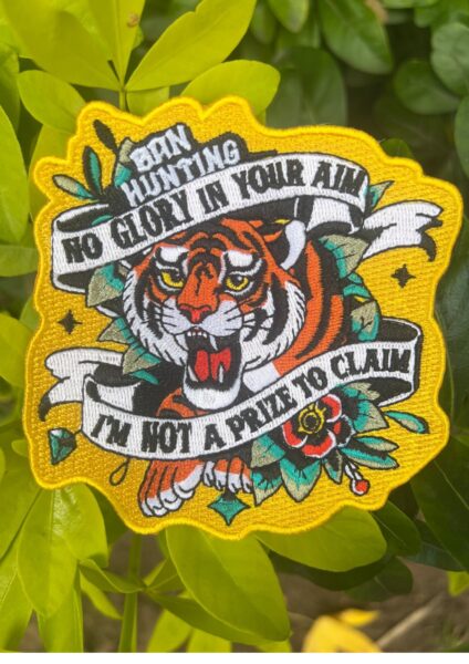 Embroidered patch featuring a roaring tiger surrounded by floral motifs and bold banners. Text reads: “Born Hunting / No Glory In Your Aim / I'm Not A Prize To Claim.” Vivid colors and yellow border emphasize themes of resistance and animal dignity.