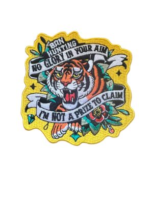 Embroidered patch featuring a roaring tiger surrounded by floral motifs and bold banners. Text reads: “Born Hunting / No Glory In Your Aim / I'm Not A Prize To Claim.” Vivid colors and yellow border emphasize themes of resistance and animal dignity.