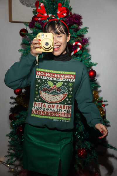 Person wearing a green organic cotton vegan Christmas jumper with humorous food-themed slogans like “HO-HO-HUMMUS” and “FALA-LA-LA-FALAFEL,” standing in front of a decorated Christmas tree and holding a yellow camera, smiling.